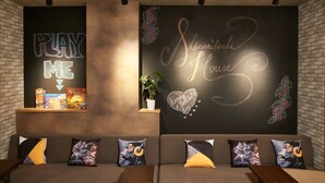 A cozy lounge area with a dark wall featuring a chalkboard with 'PLAY ME' and 'Shinsaibashi House' written on it. There are books and a plant on a shelf, and a couch with colorful cushions. - WELLSTAY 　Osaka　Shinsaibashi Apartment hotel (Osaka)