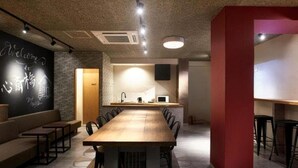 A modern dining area with a long wooden table, black chairs, and a red accent wall. - WELLSTAY　Osaka　Shinsaibashi Apartment hotel (Osaka)