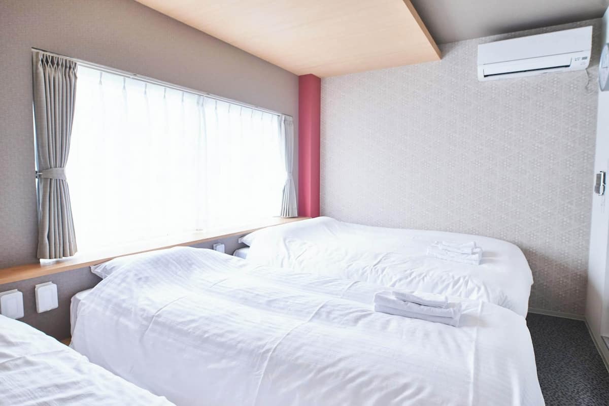 A hotel room with two beds, a window with curtains, a wooden headboard, a wall-mounted air conditioner, and a door.