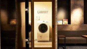 A laundry room with a washing machine, a small table, and a shelf with books. - WELLSTAY　Osaka　Shinsaibashi Apartment hotel (Osaka)