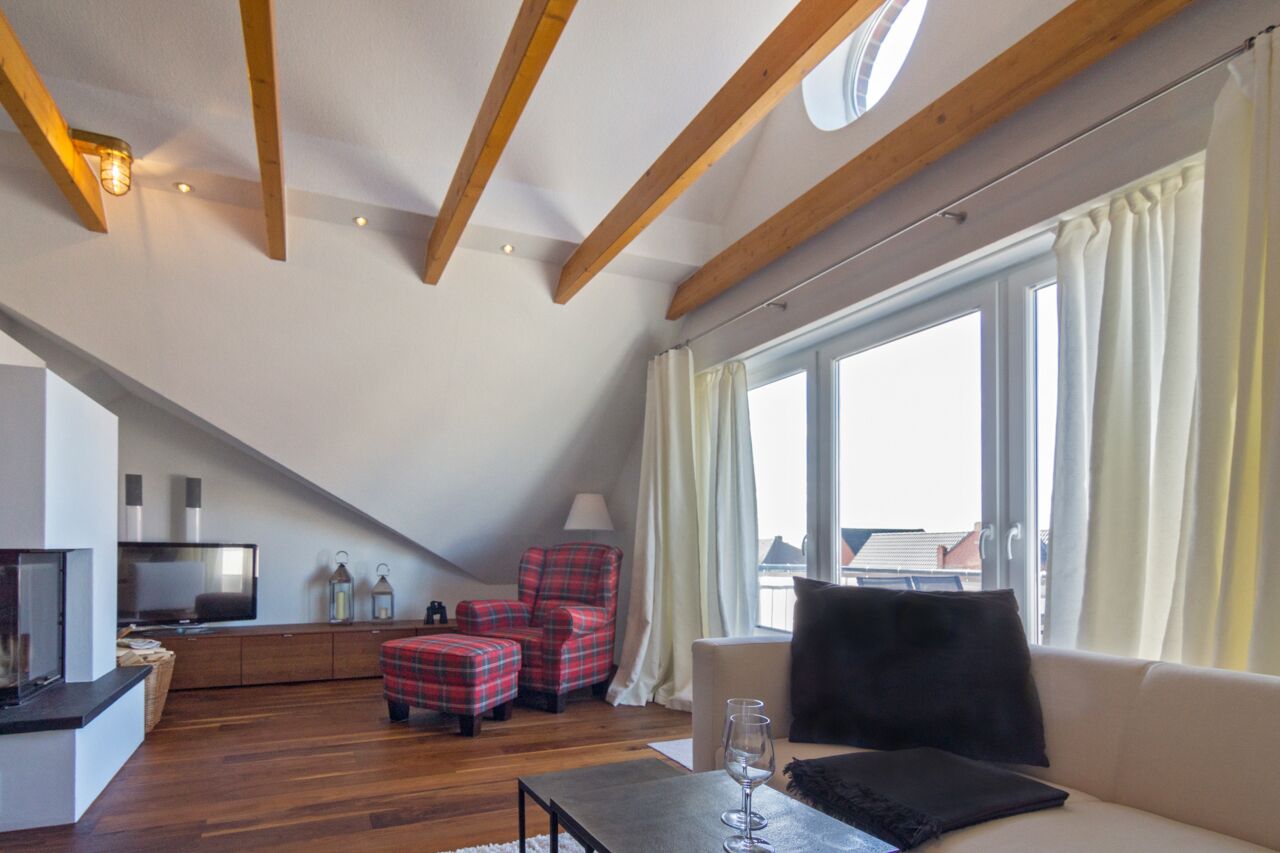 Une pièce de style « loft » avec un plafond en pente, des poutres en bois, une grande fenêtre avec des rideaux, un canapé, un fauteuil à carreaux rouges, une petite table avec un vase et un téléviseur posé sur un meuble.
