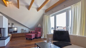 A loft-style room with a sloped ceiling, wooden beams, a large window with curtains, a sofa, a red plaid chair, a small table with a glass, and a television on a cabinet. - Vacation apartment Norddeich Loft (Norddeich)
