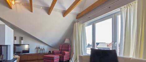 Une pièce de style « loft » avec un plafond en pente, des poutres en bois, une grande fenêtre avec des rideaux, un canapé, un fauteuil à carreaux rouges, une petite table avec un vase et un téléviseur posé sur un meuble.