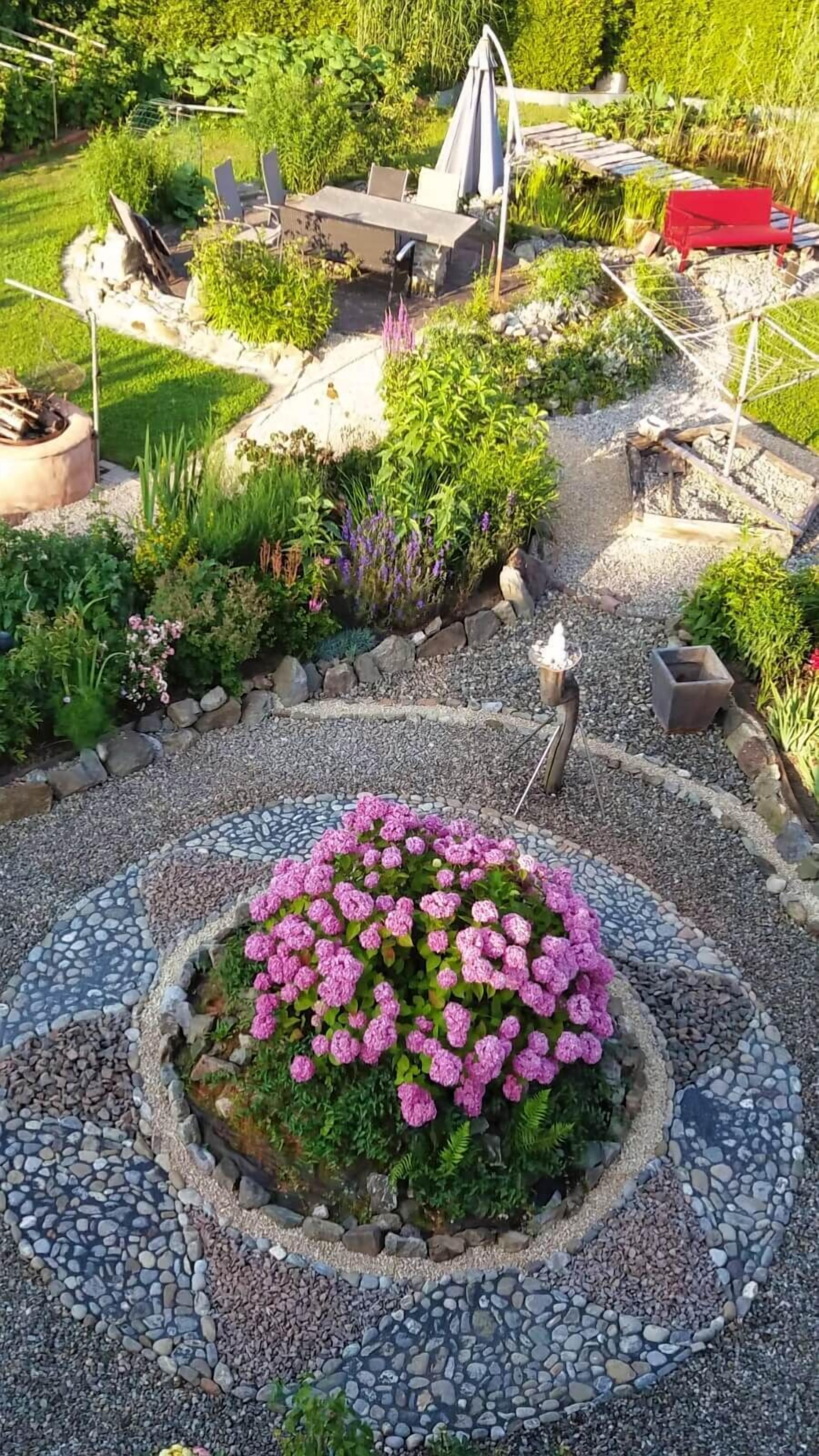 Een tuin met een rond bloembed, een vuurkorf en een zithoek met stoelen en een tafel.