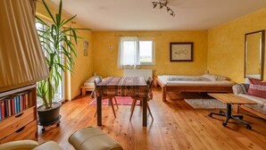Una stanza con pavimento in legno, un tavolo da pranzo con tovaglia fantasia, un letto con materasso bianco, una pianta in vaso di grandi dimensioni e una libreria piena di libri.