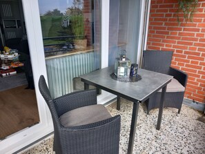 A patio area with a table and two wicker chairs, a lantern, and a view through a glass door. - Close to the beach... directly behind the dike! (Dahme)
