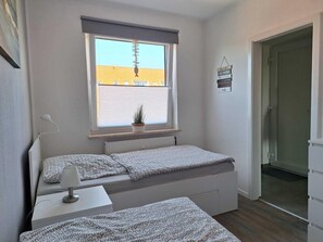 A small, well-lit room with two beds, a window with a view of a building, a bedside table with a lamp, and a door leading to another room. - Close to the beach... directly behind the dike! (Dahme)