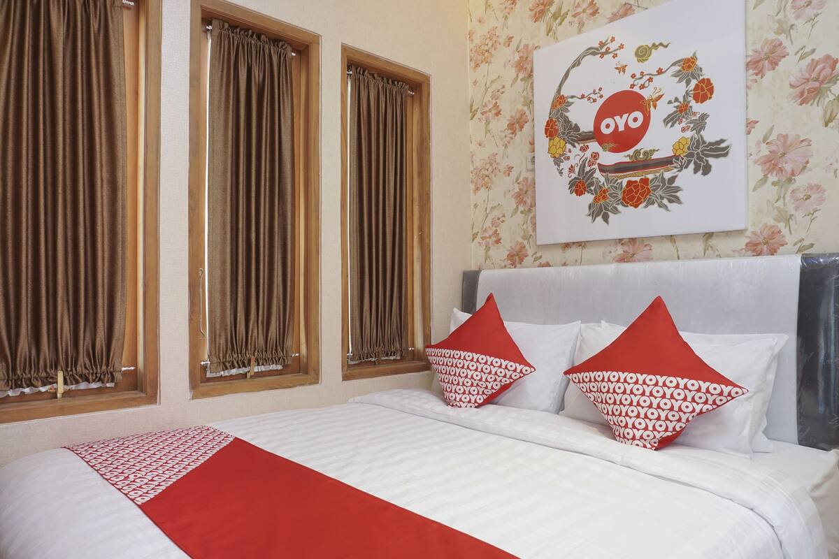A hotel room with a bed, two windows with curtains, a floral wallpaper, and a wall decoration with the word "OYO".