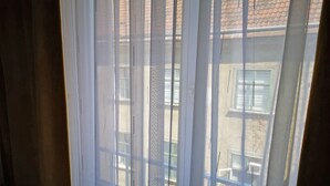 Une fenêtre avec des stores blancs, entrouverte, laissant entrevoir un bâtiment doté d’un toit en tuiles et d’un balcon.