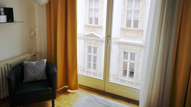 Une pièce donnant sur un bâtiment, avec une porte vitrée, un radiateur, un canapé noir orné d’un coussin à motifs et une lampe blanche fixée au mur.