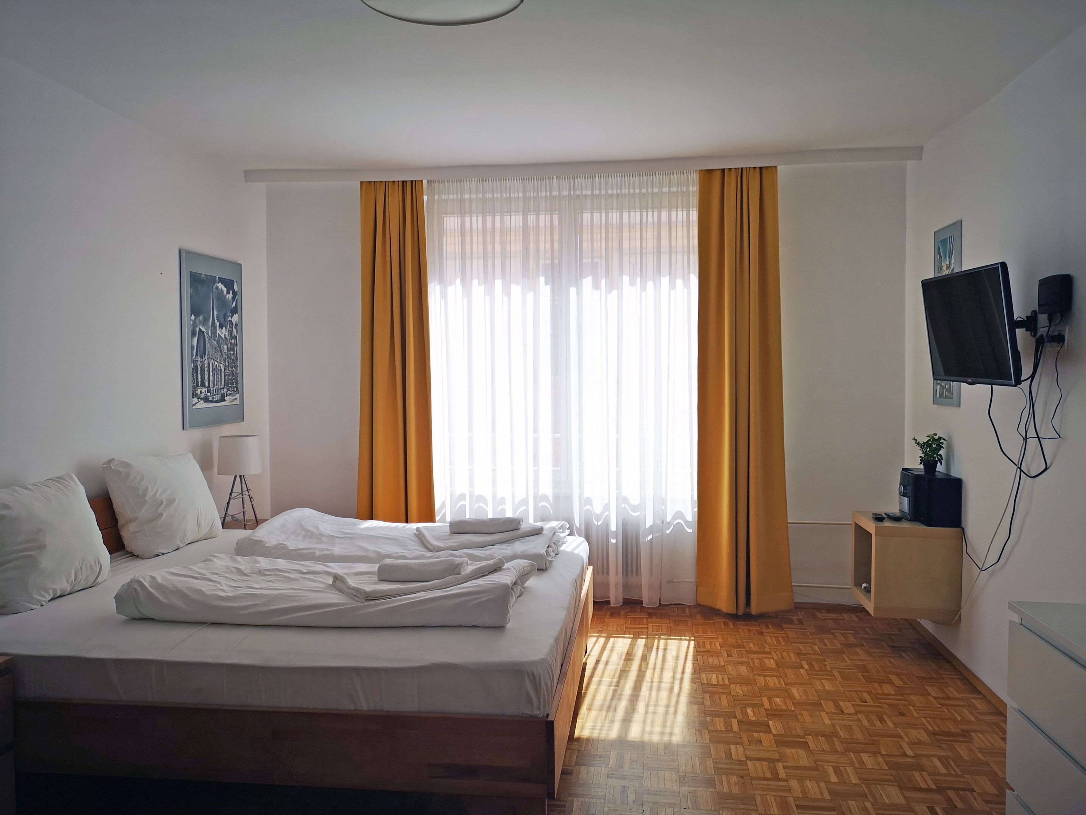 A hotel room with a bed, a bedside table, a TV mounted on the wall, and a window with curtains.