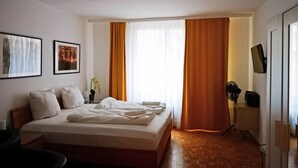 Une chambre d’hôtel comprenant un lit, un bureau, une chaise, un téléviseur fixé au mur et une fenêtre avec des rideaux.