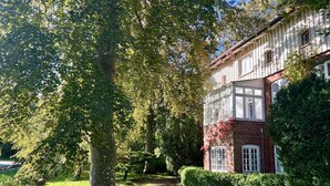 En traditionel bygning med murstensfacade, store vinduer og en velplejet have med træer og buske.