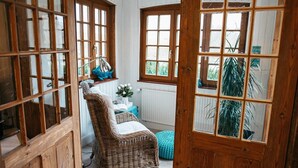 Et hyggeligt rum med en fletstol, et lille bord med en vase blomster og udsigt til grøn natur ude gennem et vindue.