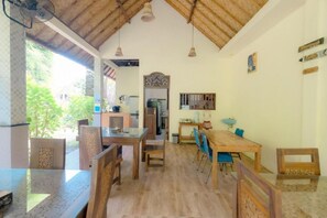Un comedor con mesas y sillas de madera, una cocina al fondo y vistas a la vegetación exterior.