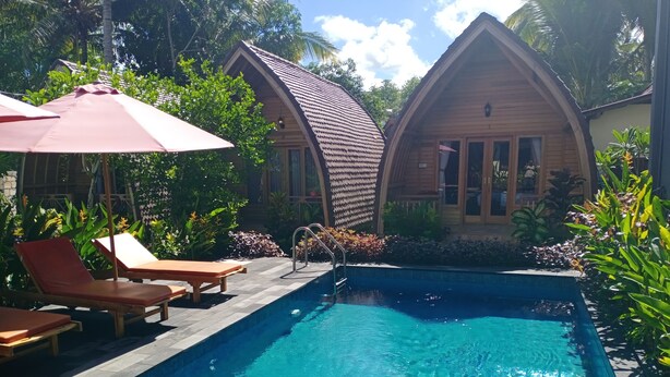 A swimming pool with lounge chairs and an umbrella, surrounded by lush greenery and wooden buildings. - Visual Inn (Penida Island)