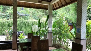 An outdoor dining area with a table and chairs, surrounded by lush greenery and a thatched roof structure. - Visual Inn (Penida Island)