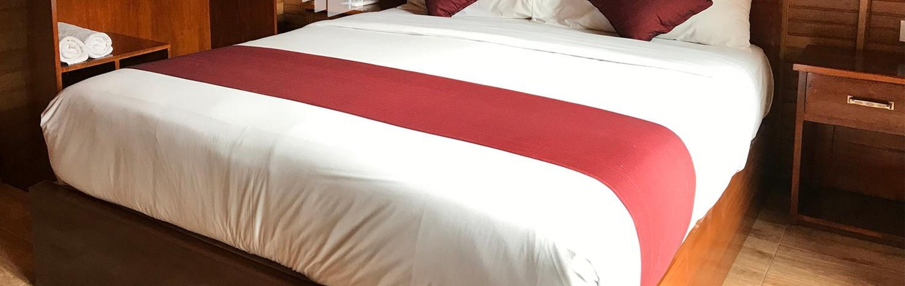 Ein Hotelzimmer mit einem ordentlich bezogenen Bett, einem Nachttisch aus Holz mit einer Lampe und einem Holzschrank.