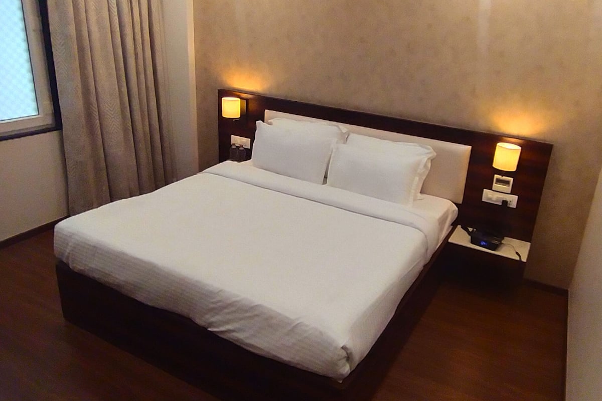 Una cama bien hecha con sábanas blancas en una habitación de hotel. Hay dos mesitas de noche, una de ellas con lámpara y reloj.
