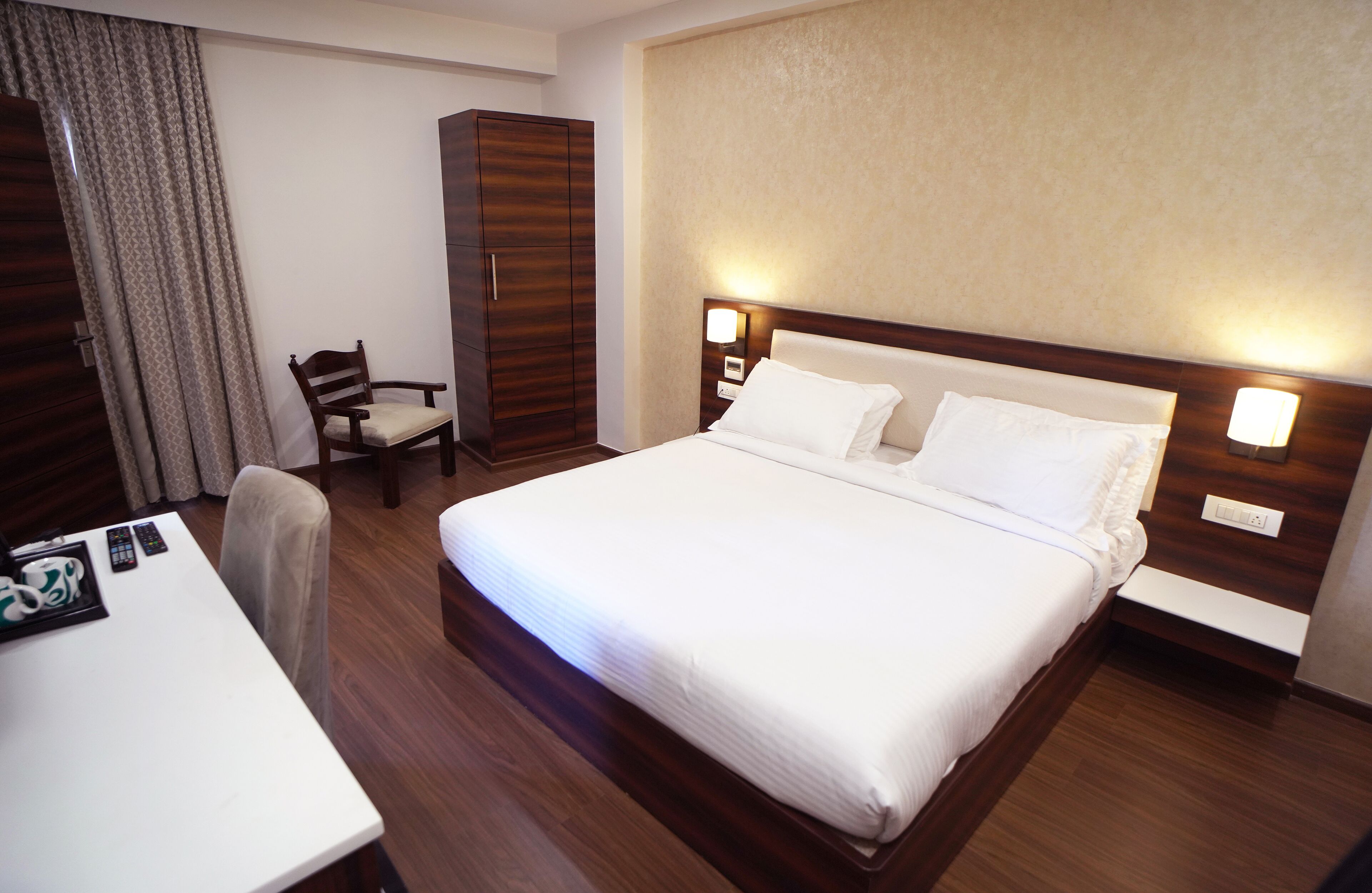 A hotel room with a large bed, a wooden wardrobe, a desk with a chair, and a nightstand with a lamp.