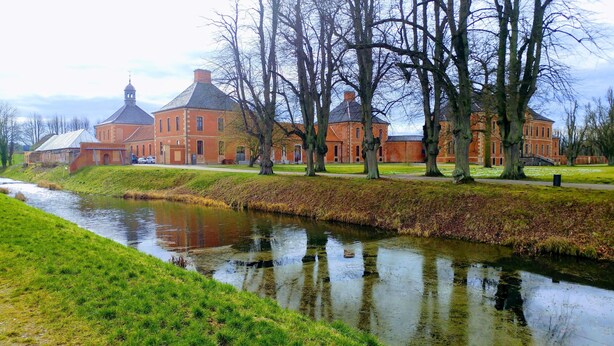 A large red brick building with a prominent tower, situated beside a calm river with a grassy bank.
