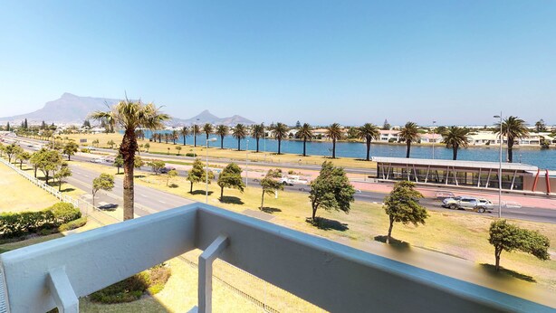 A balcony with a view of a coastal road, palm trees, and a body of water. - Palo Alto Holiday Condo Bloubergstrand (Cape Town)
