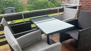 A balcony with wicker furniture, a glass table, and a view of a garden and buildings. - Robby 6 - Near the beach, quiet apartment with balcony (Wangerooge)