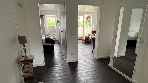 A modern living room with a dark wood floor, a sofa, a lamp, and a view into a bedroom with a bed and a window. - Robby 6 - Near the beach, quiet apartment with balcony (Wangerooge)