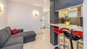 Ein modernes Wohnzimmer mit grauem Sofa, roten Kissen, einer Küchenzeile mit Steinrückwand und roten Barhocker.