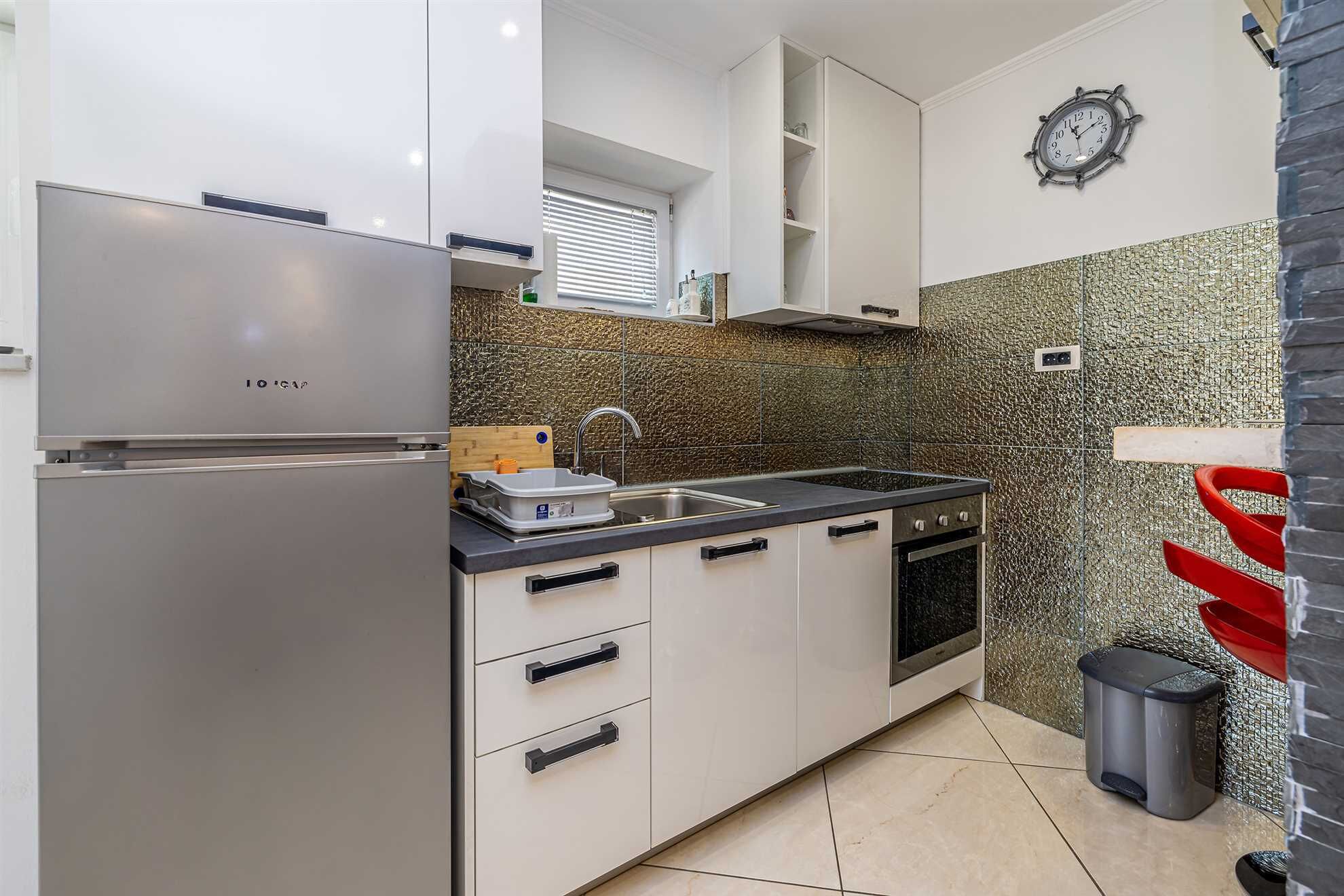 A compact kitchen with a stainless steel refrigerator, a sink, a stove, and a clock on the wall.