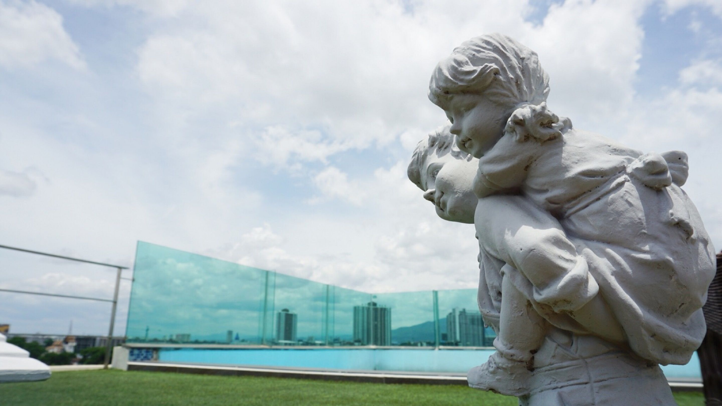 A statue of a child holding an adult's head, set against a backdrop of a modern building and a clear sky.