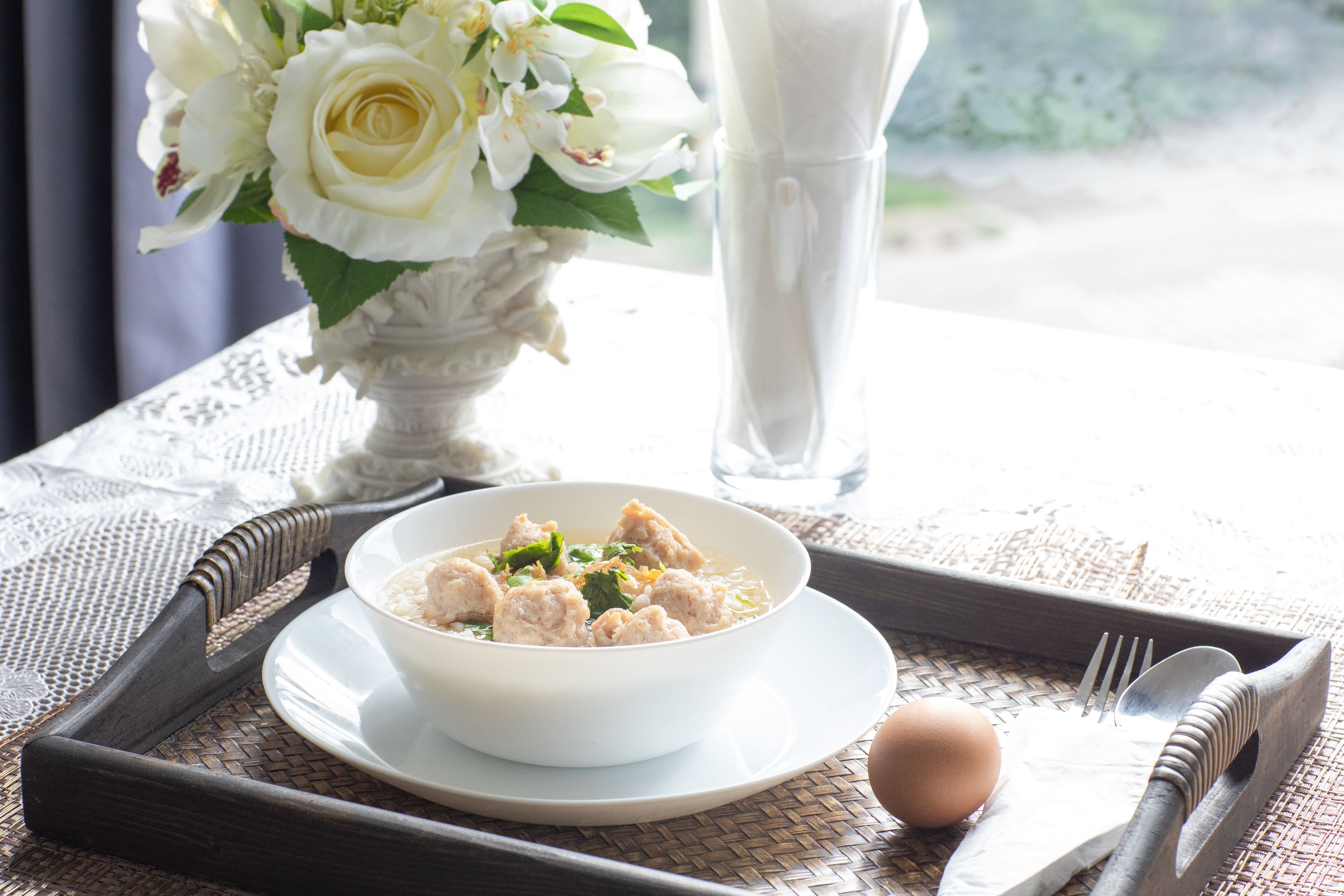 A tray with a bowl of soup, a cup of milk, a vase of flowers, and a plate with a brown egg.