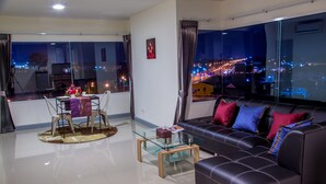 A modern living room with a glass coffee table, a black leather sofa with colorful cushions, a dining area with a table and chairs, and large windows offering a city view at night. - The Greenery Central Suites (San Sai)