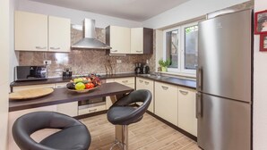 Een moderne keuken met een roestvrijstalen koelkast, een koffiezetapparaat en een fruitschaal op het aanrecht. Er staan twee zwarte barkrukken en er is een raam met uitzicht op groen.