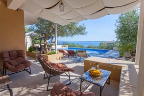 Eine sonnendurchflutete Terrasse mit Sitzgelegenheiten im Freien, einem Pool und Blick auf das Meer.