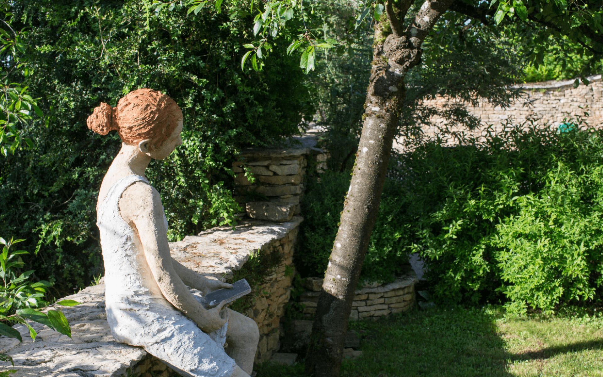 A statue of a woman seated on a stone bench in a garden with stone walls and lush greenery.