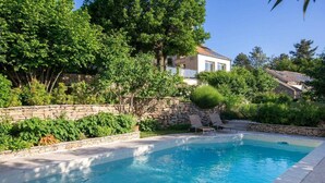Una piscina con muros de piedra, rodeada de abundante vegetación y una escalera de piedra que sube hacia una casa.