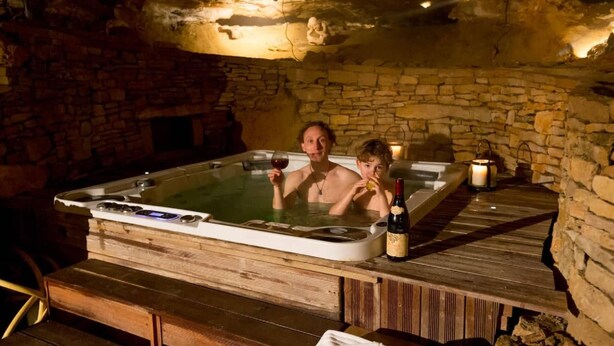 Una habitación con paredes de piedra, una bañera de madera caliente, una chimenea y una mesa con botellas y vasos.
