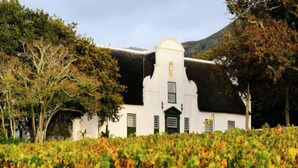 A white building with a black roof, surrounded by trees and a vineyard. - Whalesong Glencairn (Cape Town)