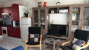 Un salon comprenant un canapé, deux fauteuils, une table basse, un téléviseur et un coin cuisine avec des placards rouges et un réfrigérateur.