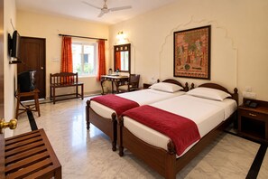 A hotel room with a bed, a desk, a chair, a television, and a painting on the wall. - WelcomHeritage Kalyan Bhawan (Jaisalmer)