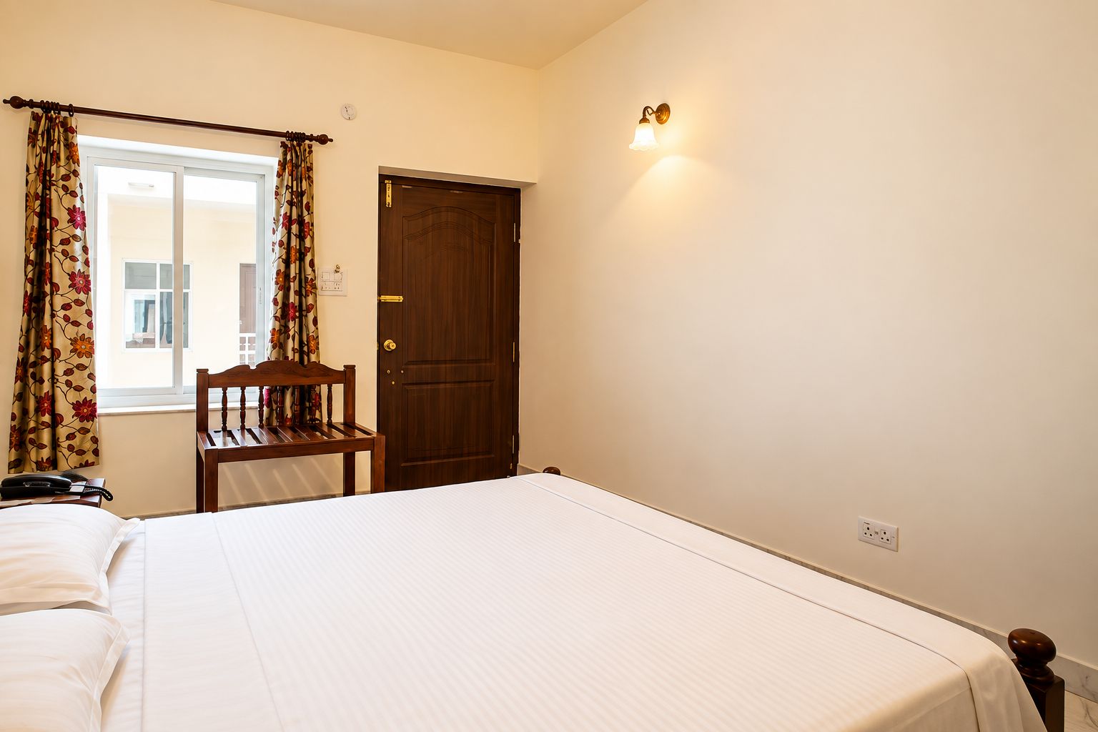 A hotel room with a bed, a chair, a wooden door, and a wall-mounted light fixture.