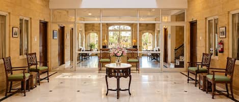 A spacious lobby with a high ceiling, a central round table with chairs, and a view of the outdoor area through large windows.