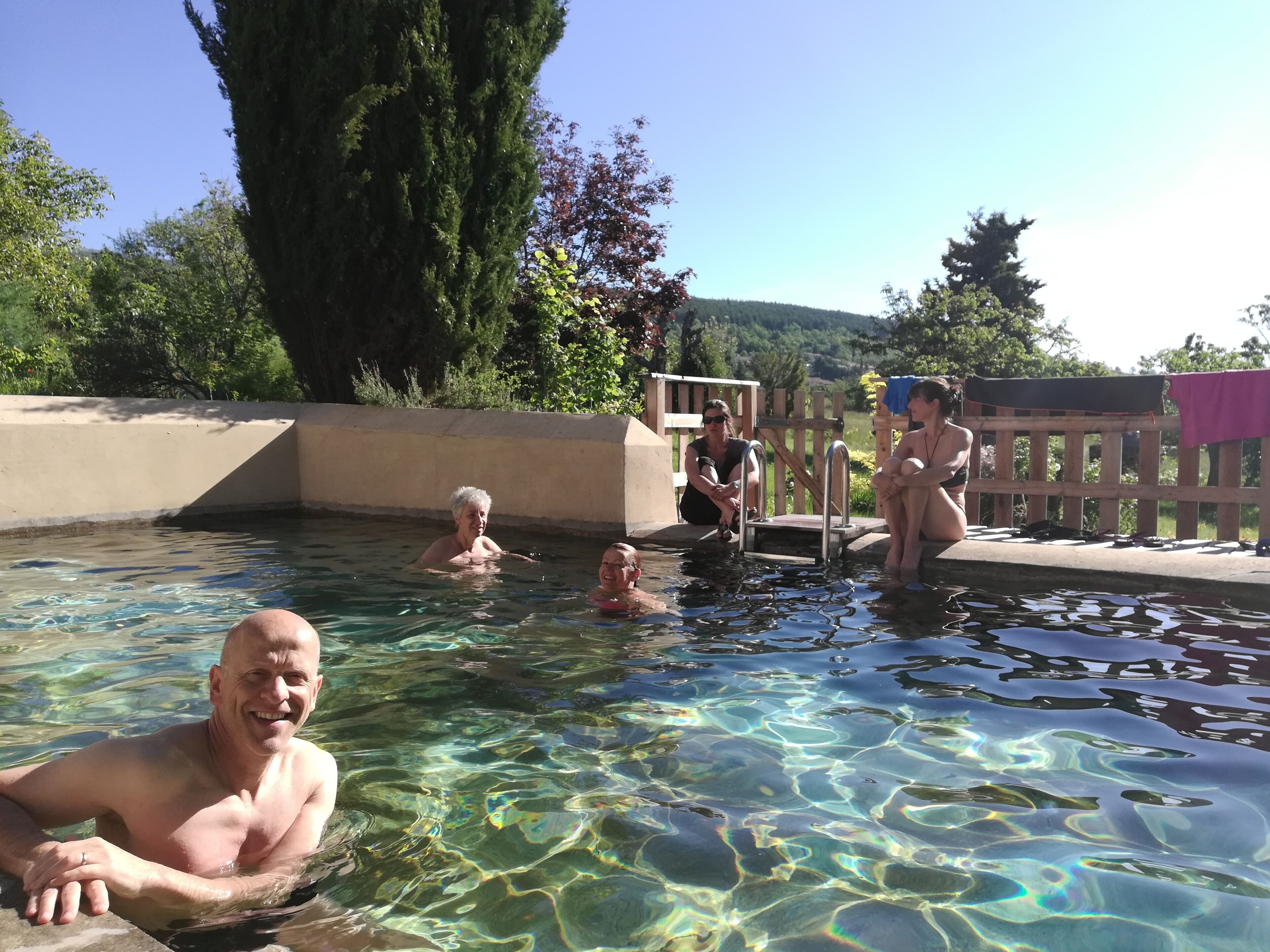 Une piscine extérieure aux eaux claires, où l’on voit des personnes se baigner, entourée d’une terrasse en bois et de verdure.