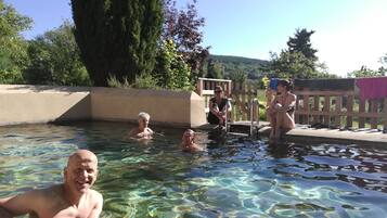 Ein klarer Außenpool mit Schwimmern, umgeben von einer Holzterrasse und viel Grün.
