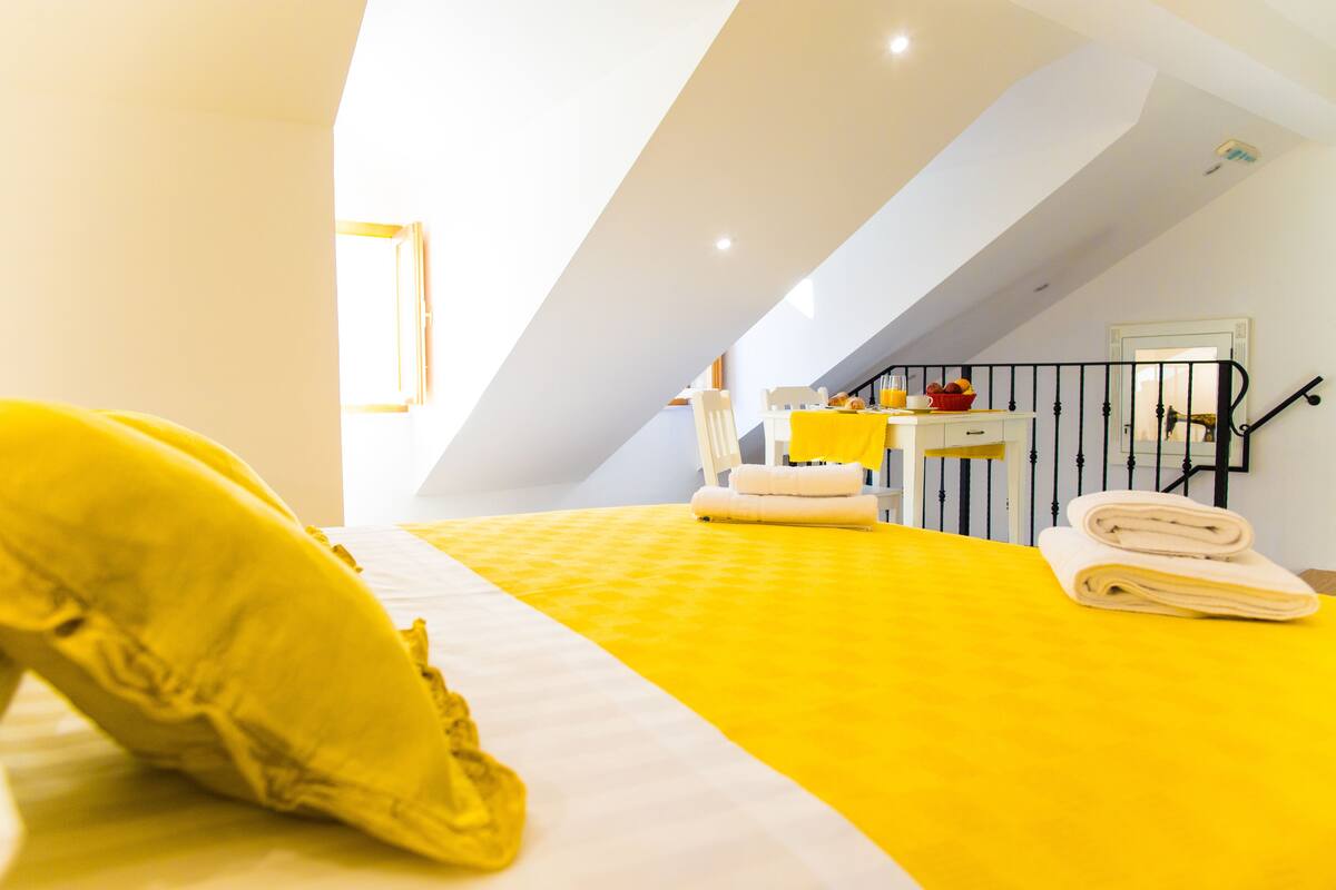 Une chambre d’hôtel avec un lit, une chaise, une petite table sur laquelle se trouve un bol de fruits, et une fenêtre.