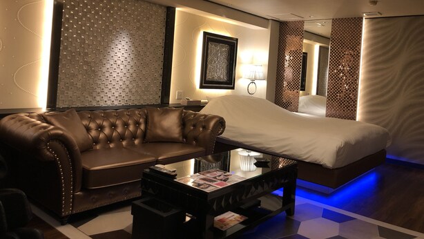 Quarto de hotel moderno com uma cama grande, um sofá de couro, uma mesa de centro de vidro e uma parede com padrão geométrico.