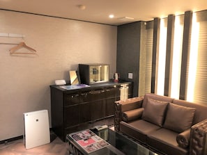 Una sala moderna con un sofá, una mesa de centro, un refrigerador pequeño, un mueble con espejo y una ventana con persianas.