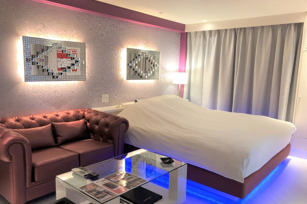 A modern hotel room with a large bed, a leather sofa, a glass coffee table, and decorative wall mirrors.