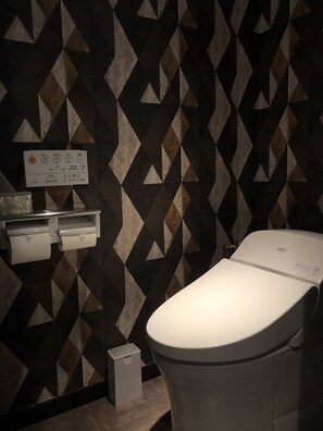 Un baño moderno con pared de diseño geométrico, inodoro, dispensador de toallas de papel y un cesto de basura.
